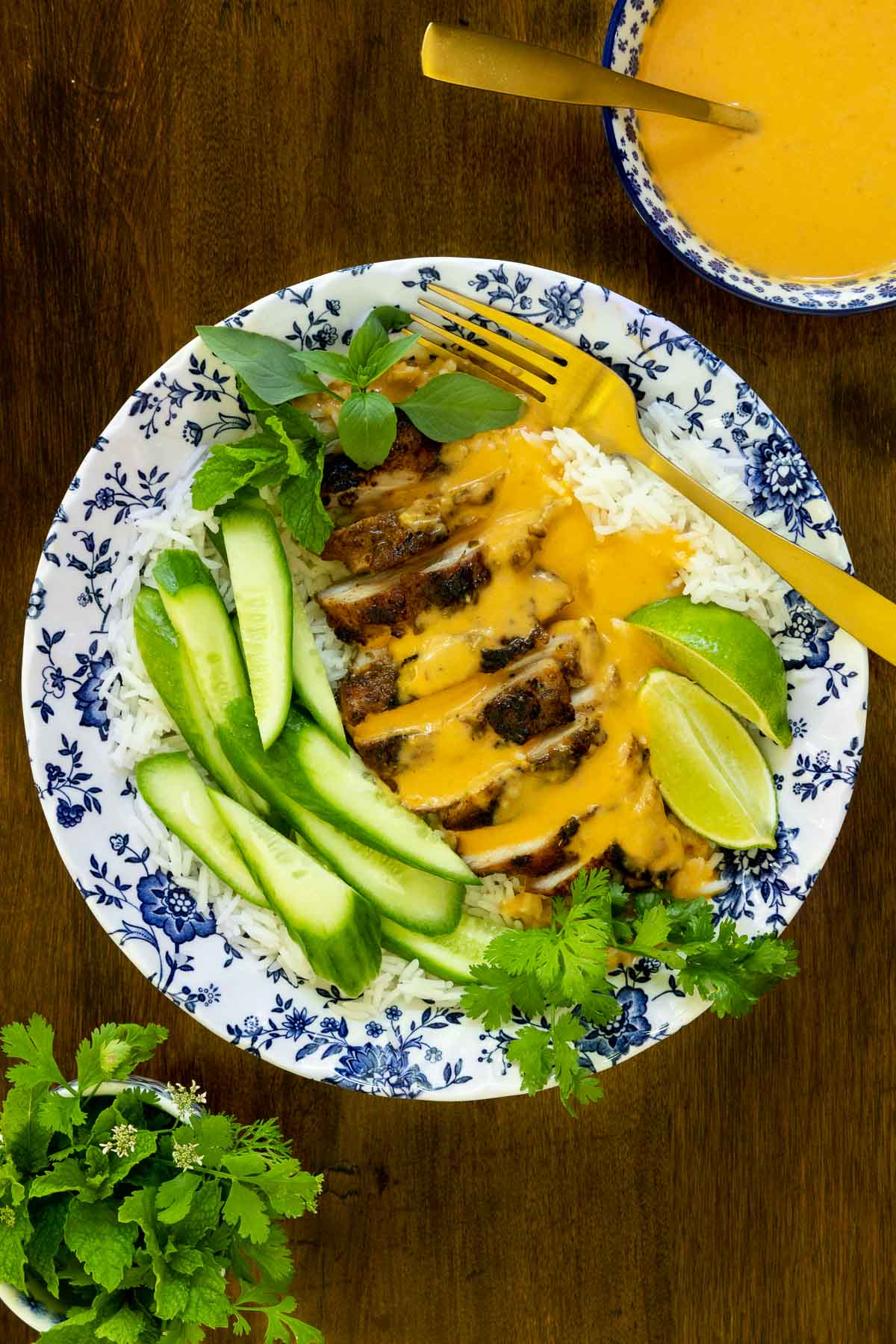 Vertical overhead picture of Grilled Chicken Thighs with Coconut Chili Lime Sauce in a blue and white bowl with cucumbers and herbs
