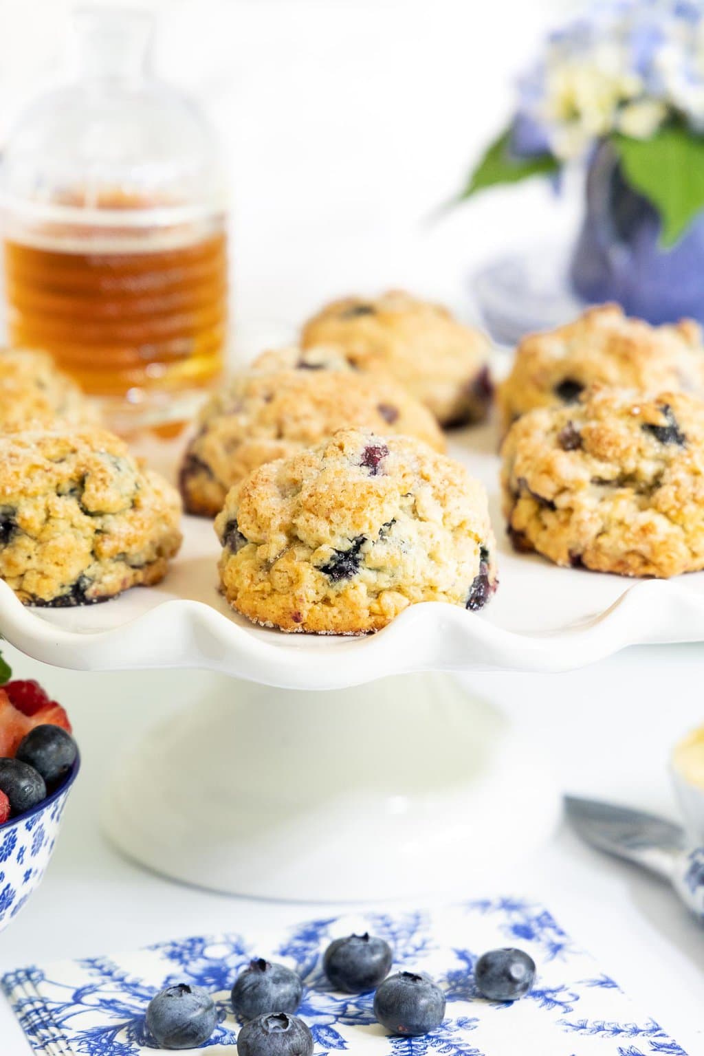Vertical picture of blueberry scones on a white cake stand with a jar of honey and flowers in the background