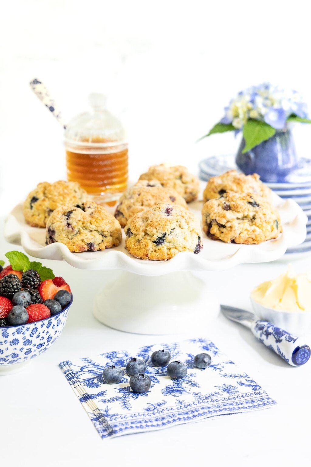 Vertical photo of a batch of Lemon Blueberry Scones on a scalloped pedestal serving plate surrounded by blueberries and other fruit.