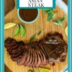 Vertical overhead photo of a Steve's Grilled Asian Steak on a round wood cutting board with fresh herbs, lime wedges and a bowl of extra sauce.