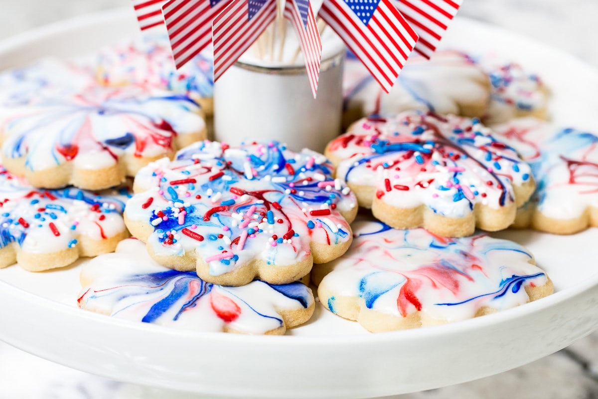 Horizontal closeup photo of a batch of Red, White and Blue Glazed Shortbread Cookies on a white serving plate with a circle of U.S. flags in the center.