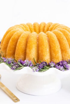 Horizontal photo of a Ridiculously Easy Sugar-Glazed Ricotta Bundt Cake on a white scalloped pedestal cake stand surrounded by fresh rosemary and lavender.