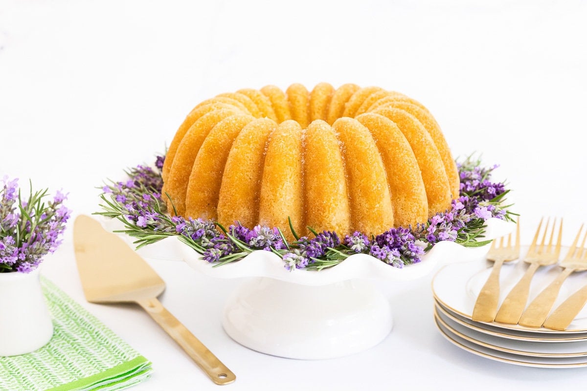 Horizontal photo of a Ridiculously Easy Sugar-Glazed Ricotta Bundt Cake on a white scalloped pedestal plate surrounded by rosemary and lavender.
