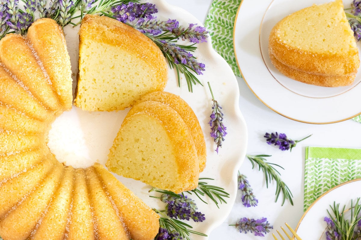 Overhead horizontal photo of a Ridiculously Easy Sugar-Glazed Ricotta Bundt Cake on a white scalloped serving plate surrounded by fresh rosemary and lavender sprigs.