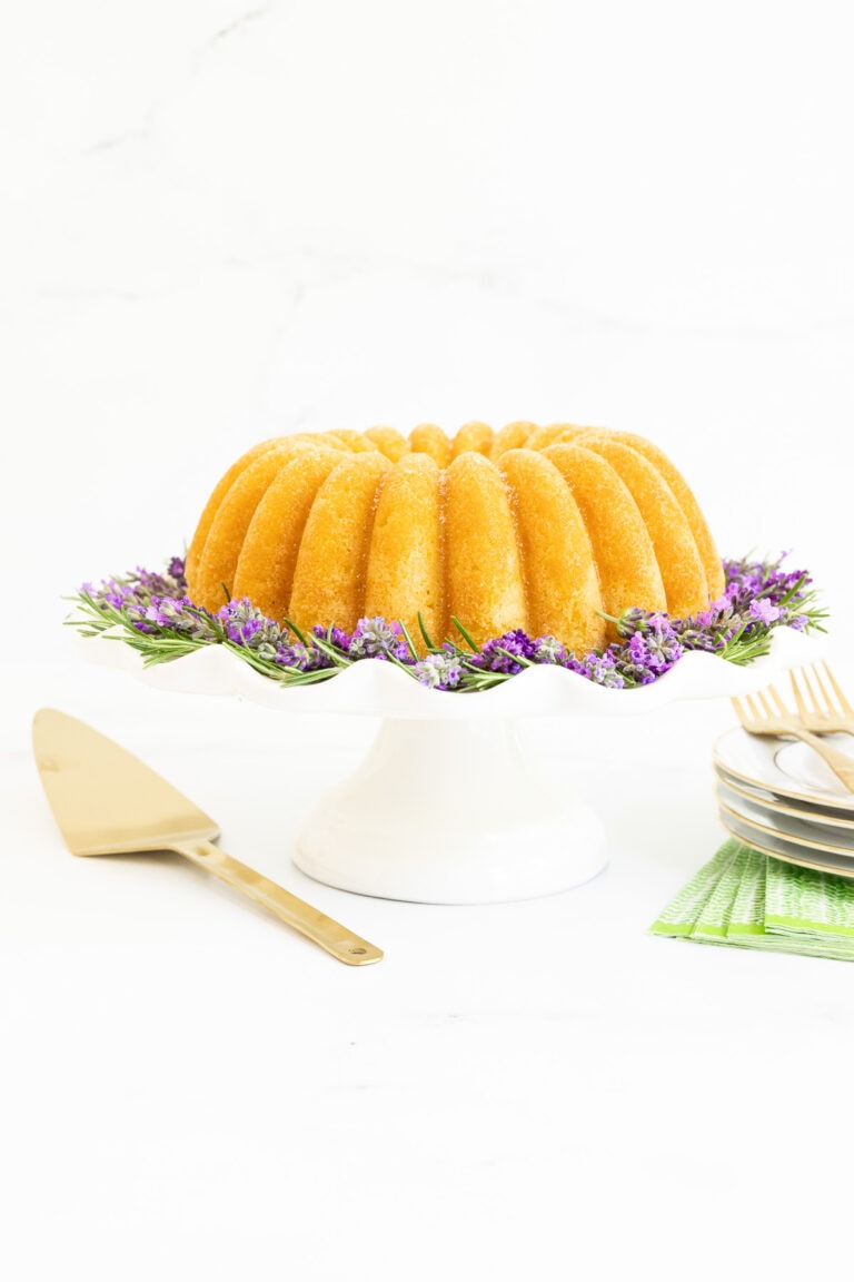 Vertical photo of a Ridiculously Easy Sugar-Glazed Ricotta Bundt Cake on a scalloped white pedestal cake stand decorated with fresh flowers.