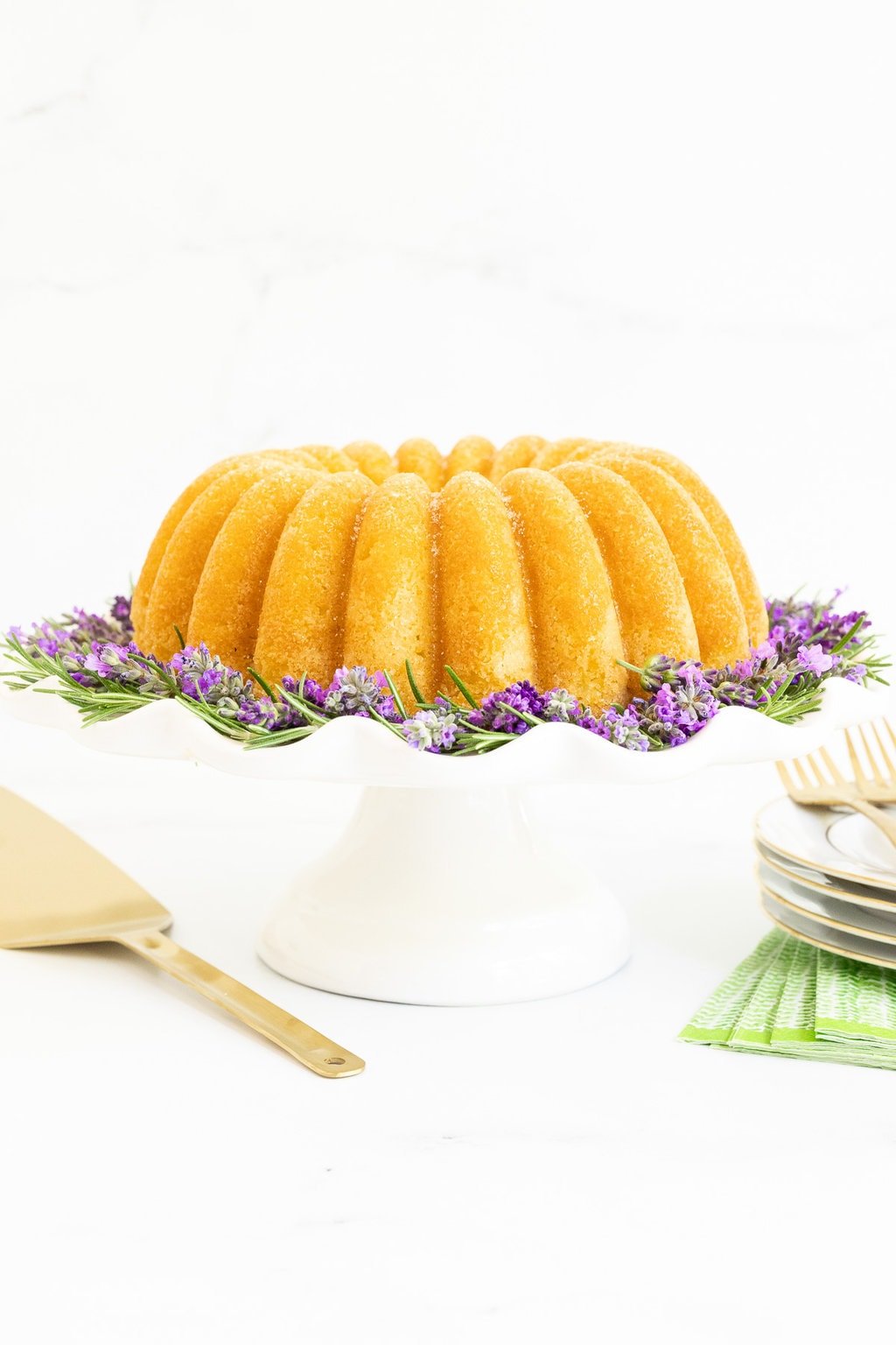 Horizontal photo of a Ridiculously Easy Sugar-Glazed Ricotta Bundt Cake on a white scalloped pedestal plate surrounded by fresh rosemary and lavender sprigs.