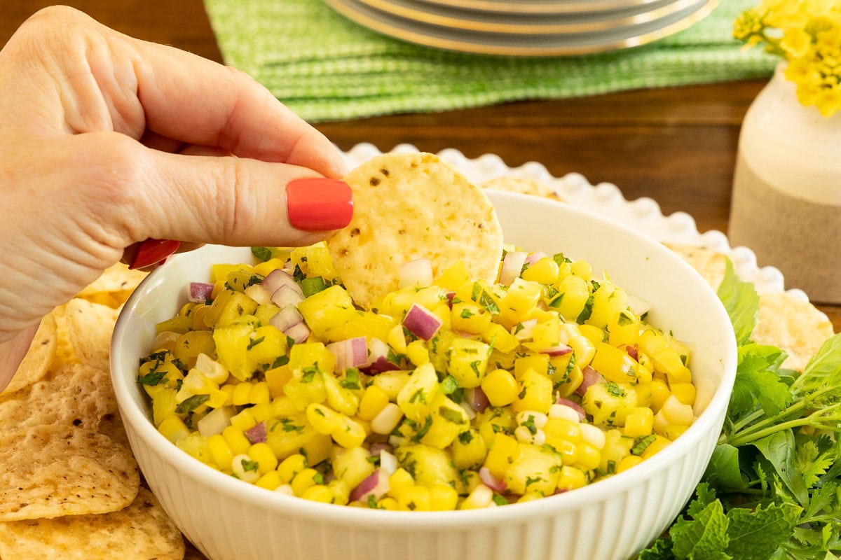 Horizontal extreme closeup photo of a hand dipping a tortilla chip into a bowl of Asian Pineapple Fresh Corn Salsa.