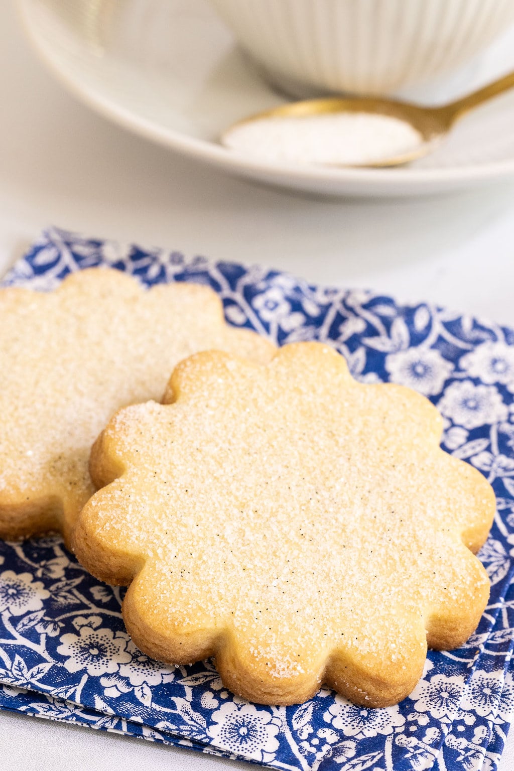Vertical closeup photo of two French Shortbread Cookies (Sablés Bretons) on a blue and white patterned napkin with a cup of tea in the background.