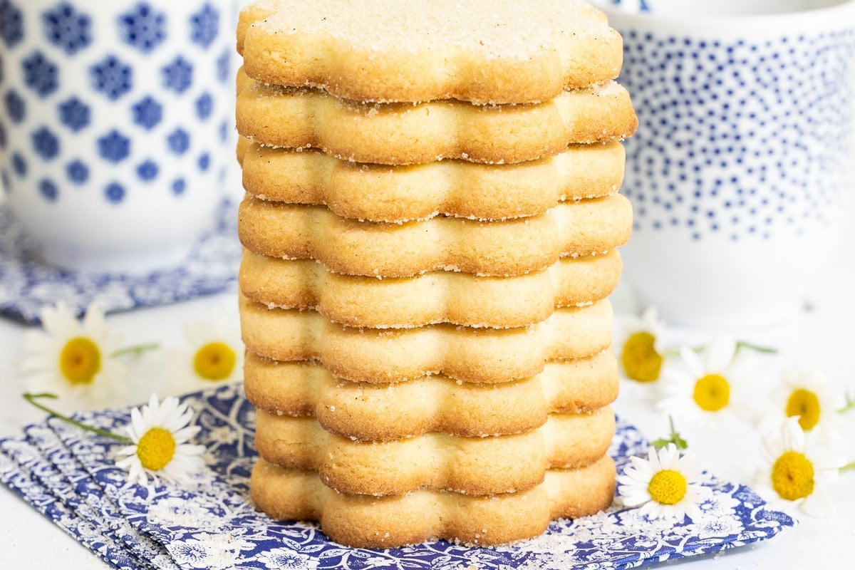 Horizontal extreme closeup photo of a stack of French Shortbread Cookies (Sablés Bretons) surrounded by flowers with cups of milk in the background.