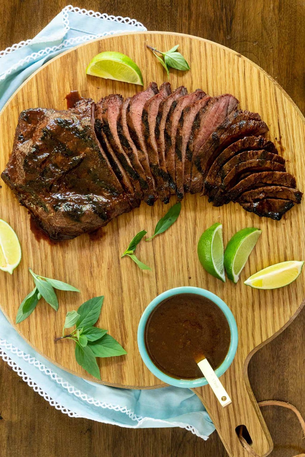 Vertical overhead photo of Steve's Grilled Asian Steak sliced on a wood cutting board with fresh herbs and lime wedges.