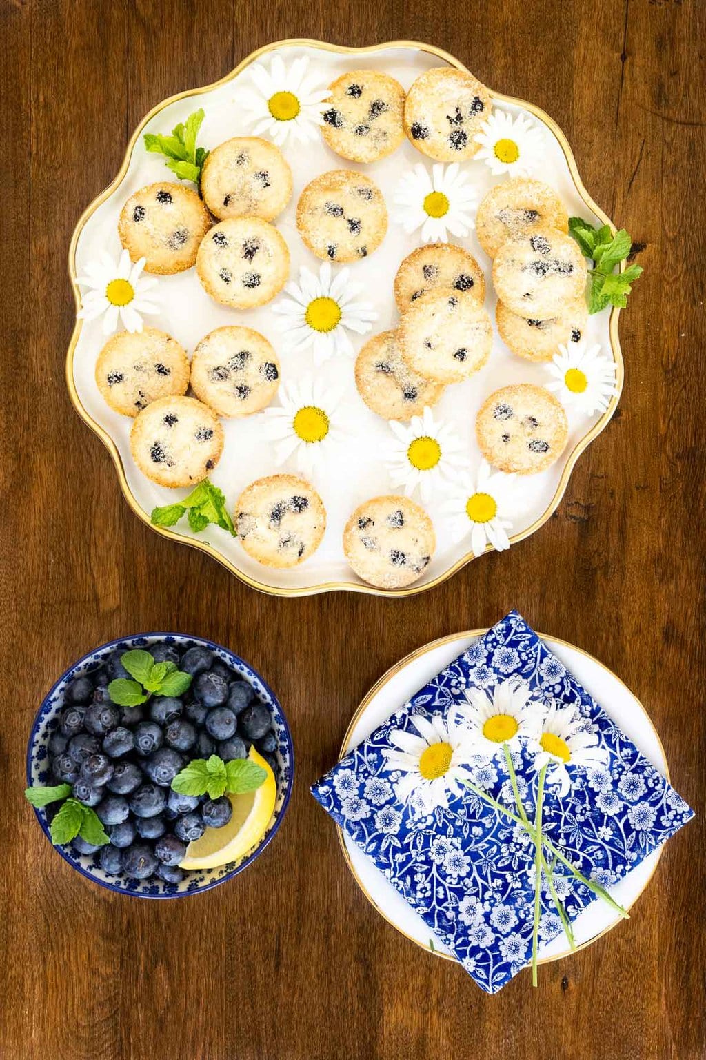 Vertical overhead photo of a plate of Ridiculously Easy Lemon Blueberry Shortbread Bites with a bowl of fresh blueberries and lemons on a wood table.