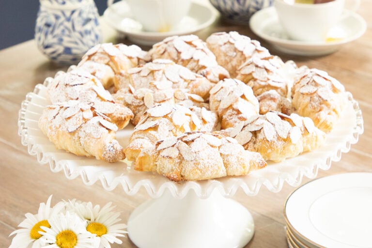Horizontal closeup photo of a batch of Ridiculously Easy Almond Croissants on a white scalloped serving pedestal plate.