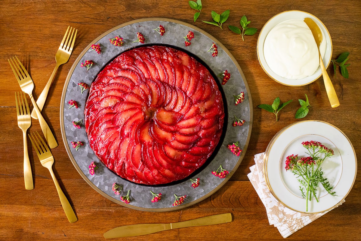 Horizontal overhead photo of a Upside Down Plum Yogurt Cake on a wood table decorated with fresh flowers and mint leaves.