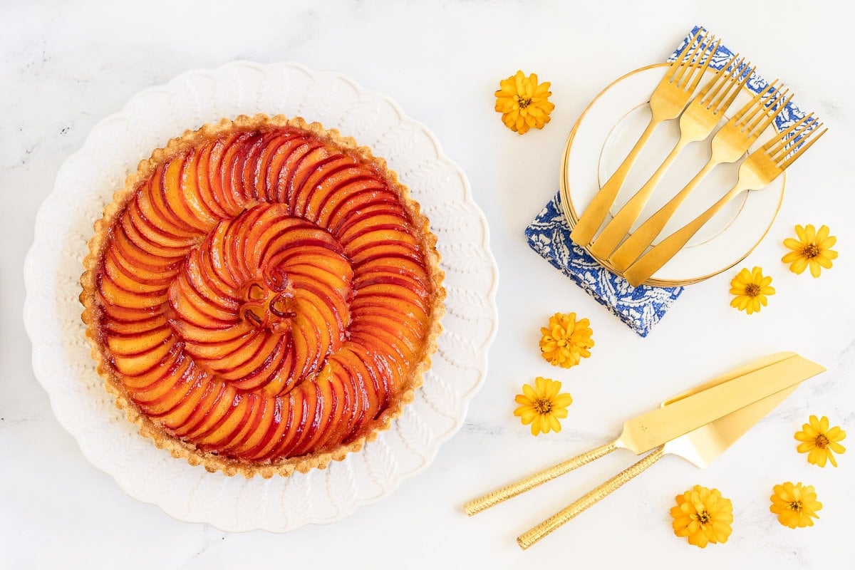Horizontal overhead photo of a French Almond Plum Tart (Tarte Et Creme D'amande) on a white serving plate surrounded by flowers and gold dinnerware.