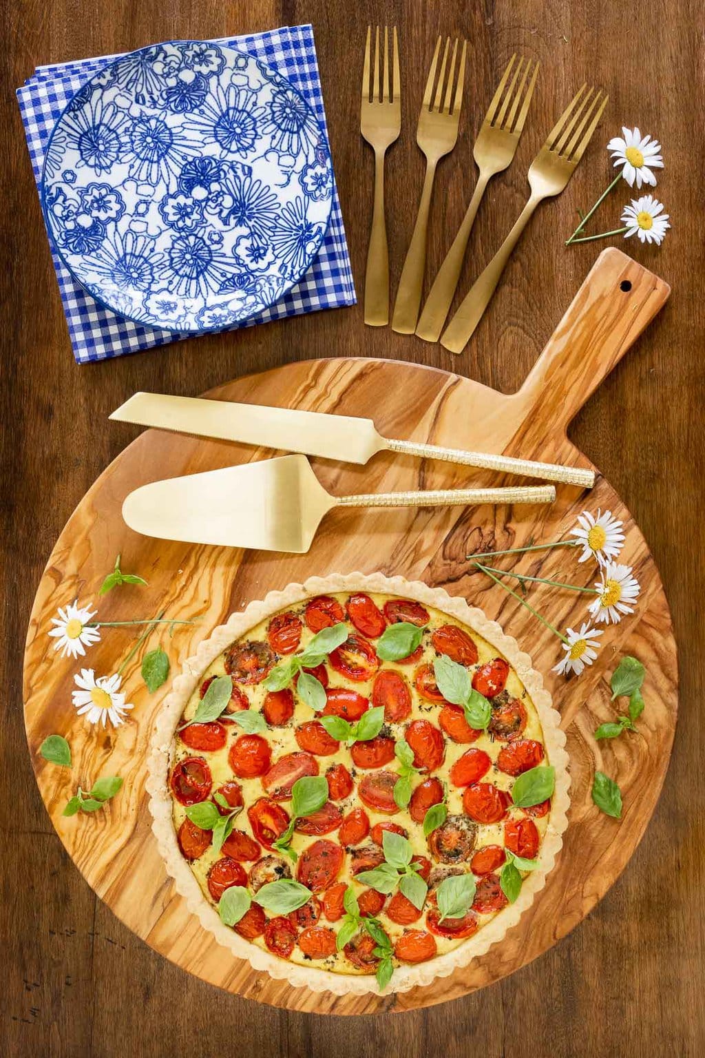Vertical overhead photo of a French Tomato Ricotta Tart on a round wood cutting board surrounded by fresh basil leaves and daisies.