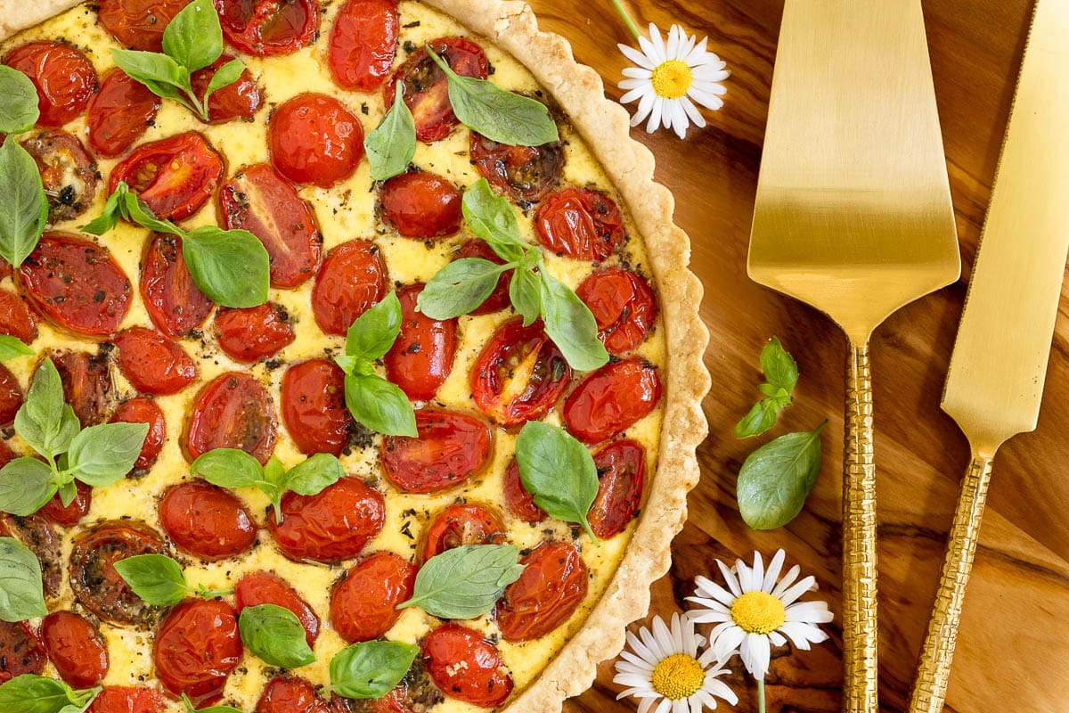 Horizontal overhead closeup photo of a French Tomato Ricotta Tart on a wood surface surrounded by daisies and fresh basil leaves.