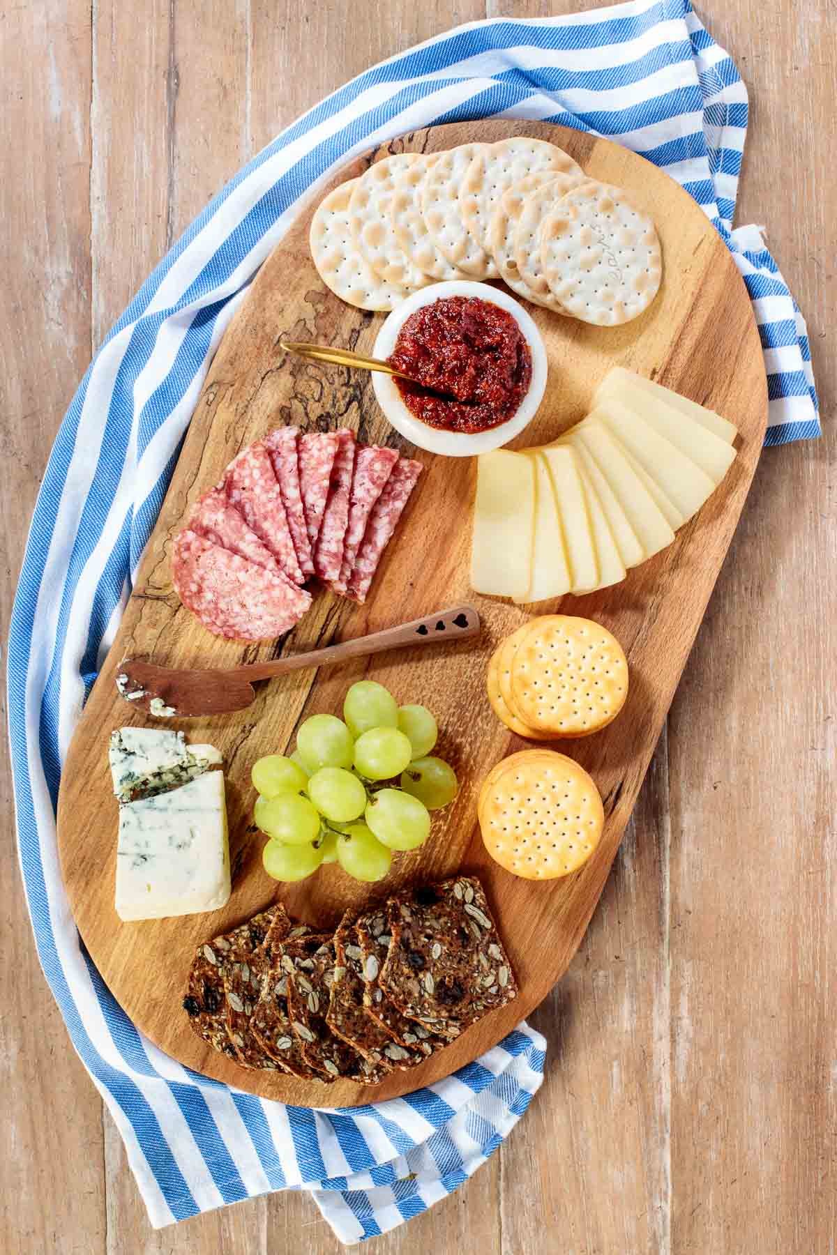 Vertical overhead photo of a charcuterie board featuring Sweet and Smoky Tomato Jam with crackers, cheeses fresh grapes and sliced sausage.