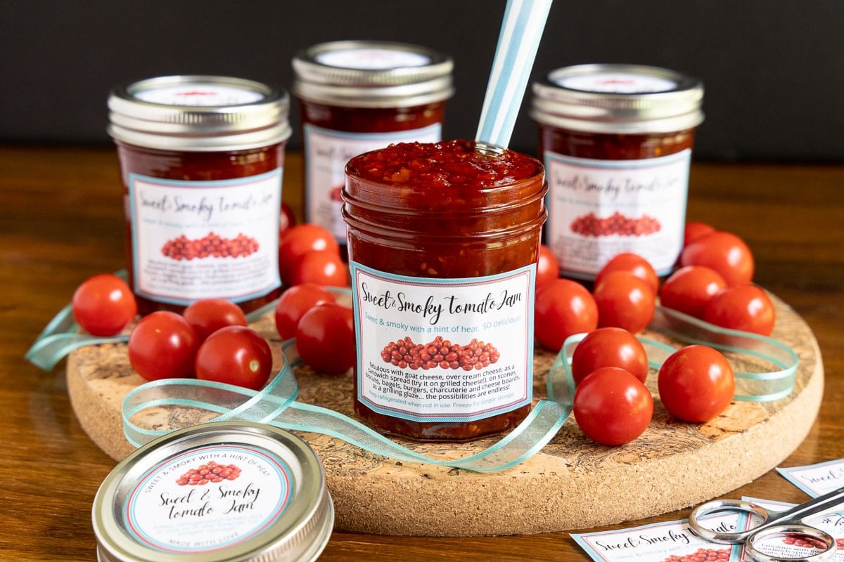 Horizontal photo of jars of Sweet and Smoky Tomato Jam surrounded by fresh cherry tomatoes. Custom gift labels are on the jars.