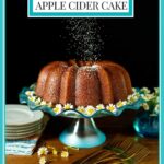 Vertical photo of a Ridiculously Easy Apple Cider Bundt Cake on a turquoise glass pedestal cake stand surrounded by flowers and with powdered sugar being sprinkled over the top of the cake.