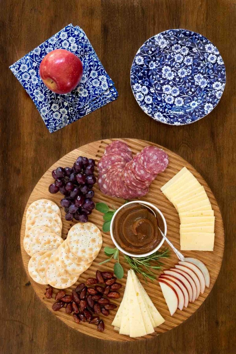 Slow cooked apple butter in a small white dish on a round board with cheese, crackers, fruit and salami.