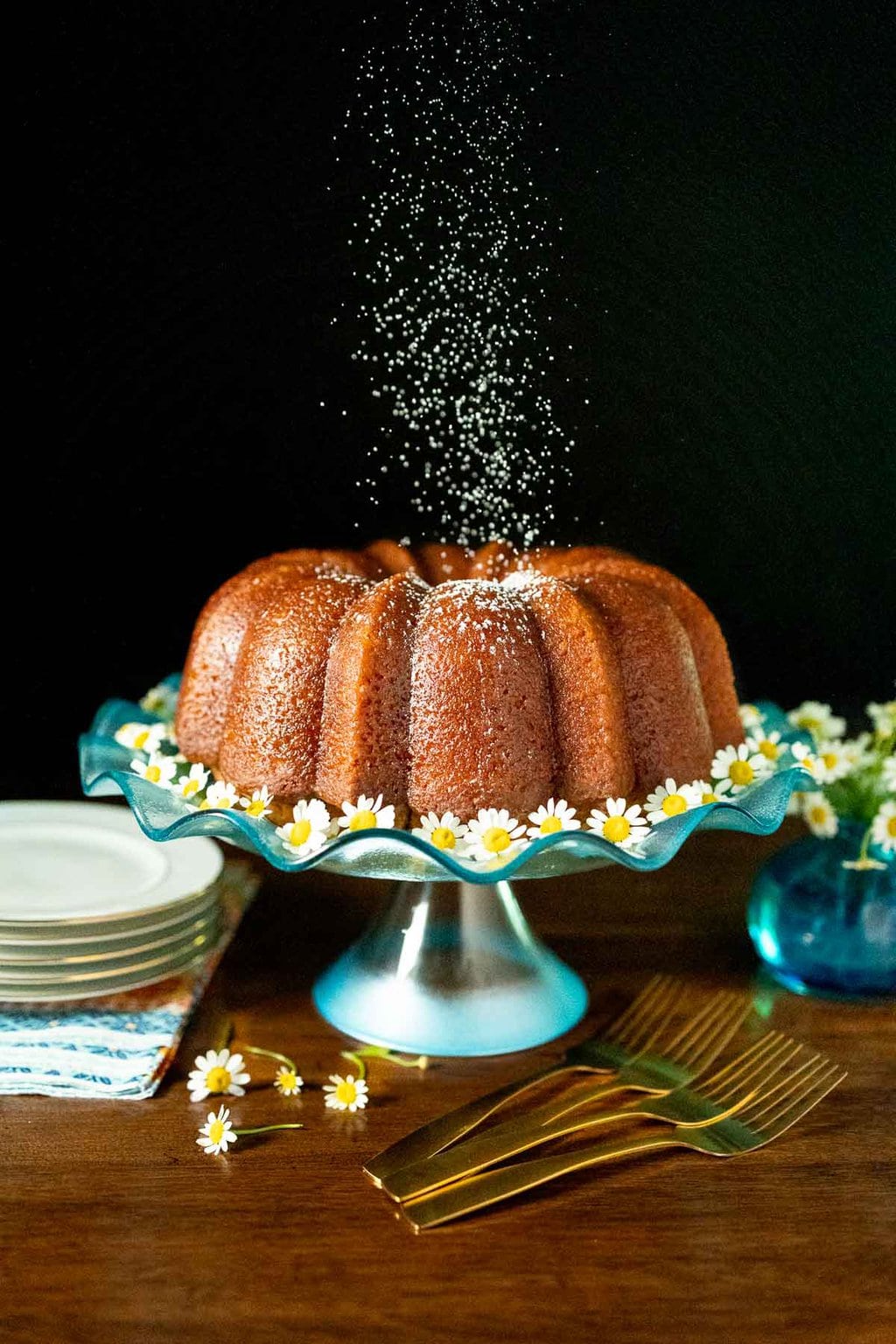 Vertical photo of a Ridiculously Easy Apple Cider Bundt Cake on a turquoise glass pedestal cake stand with powdered sugar being sprinkled over the top in a freeze action shot.