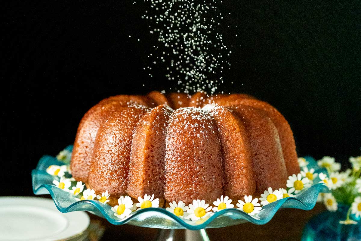 Horizontal photo of a Ridiculously Easy Apple Cider Bundt Cake on a turquoise glass pedestal cake stand surrounded with flowers and being sprinkled with powdered sugar.
