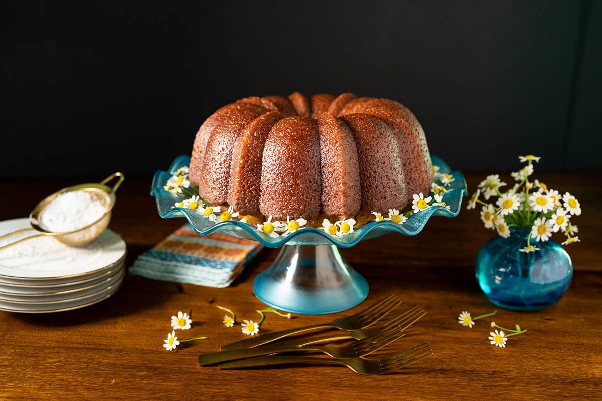 Horizontal photo of a Ridiculously Easy Apple Cider Bundt Cake on a turquoise glass pedestal stand surrounded by flowers.