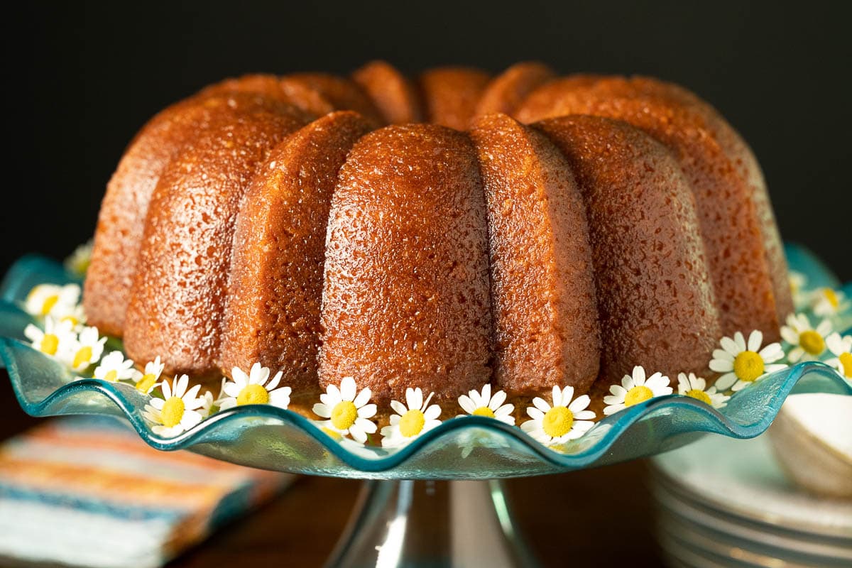 Horizontal closeup photo of a Ridiculously Easy Apple Cider Bundt Cake on a turquoise glass pedestal cake stand surrounded by flowers.