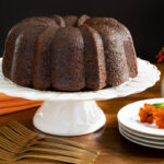 Horizontal photo of a Ridiculously Easy Butterscotch Glazed Pumpkin Bundt Cake on a white pedestal cake stand with orange flowers and napkins in the background.