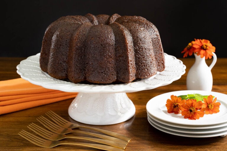 Horizontal photo of a Ridiculously Easy Butterscotch Glazed Pumpkin Bundt Cake on a white pedestal cake stand with orange flowers and napkins in the background.