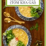 Vertical overhead photo of two serving bowls of Thai Coconut Chicken Soup (Tom Kha Gai) garnished with rice, fresh vegetables, Serrano peppers, herbs and lime wedges.