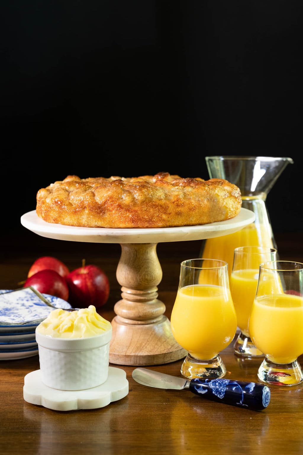 Vertical photo of a Ridiculously Easy Apple Cinnamon Breakfast Focaccia on a pedestal serving plate surrounded by fresh apples and cups of orange juice.