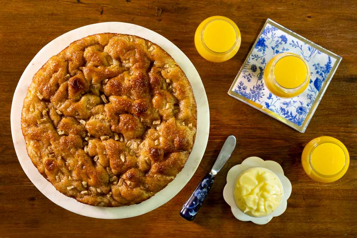 Horizontal overhead photo of a Ridiculously Easy Apple Cinnamon Breakfast Focaccia surrounded by glasses of orange juice and a cup of fresh butter on a wood table.