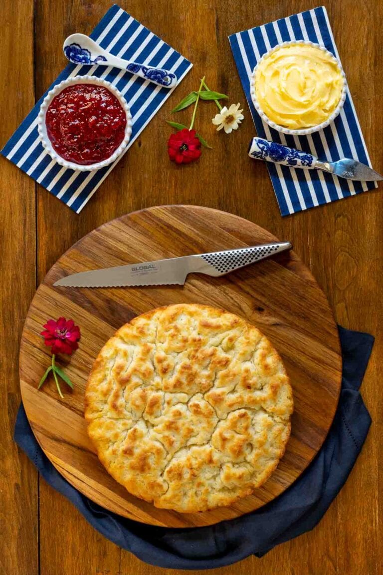 Biscuit focaccia bread on a round wooden cutting board with small dishes of butter and jam on blue and white stripped napkins.