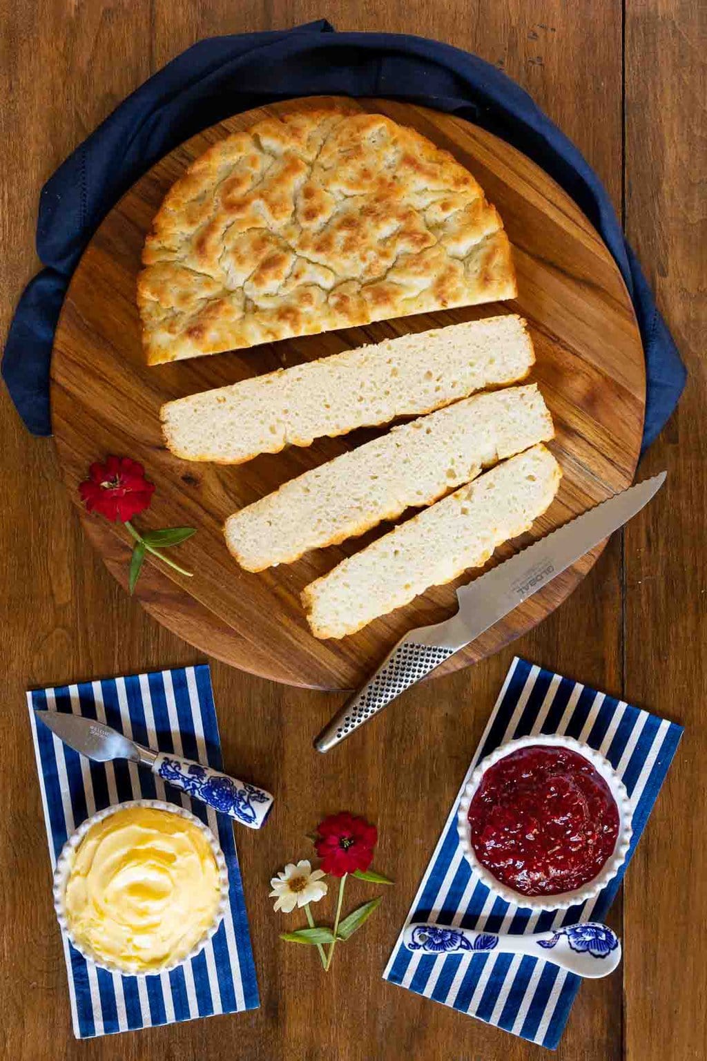 Vertical overhead photo of a sliced loaf of Ridiculously Easy Biscuit Focaccia on a round wood cutting board with bowls of raspberry jam and fresh butter.