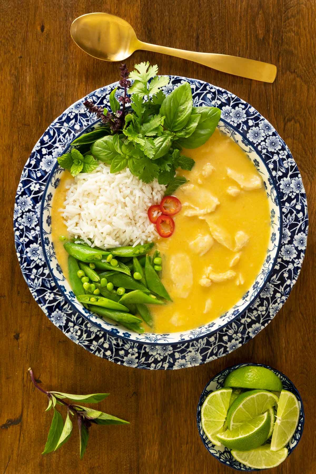 Vertical overhead photo of a bowl of Thai Coconut Chicken Soup on a wood table with rice, fresh vegetables, herbs, lime wedges and Serrano peppers.
