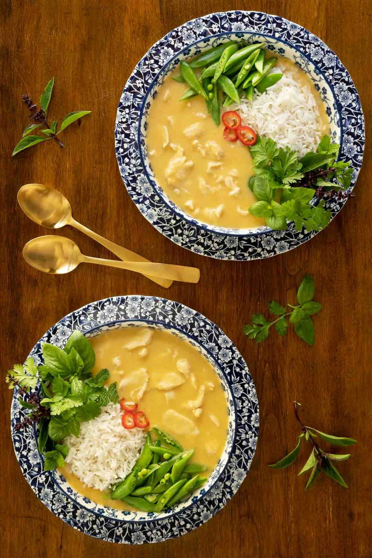 Vertical overhead photo of two serving bowls of Thai Coconut Chicken Soup garnished with rice, fresh vegetables, peppers and herbs.