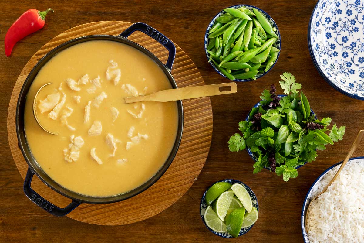 Horizontal overhead photo of a pot of Thai Coconut Chicken Soup surrounded by small bowls of fresh vegetables and herbs on a wood table.