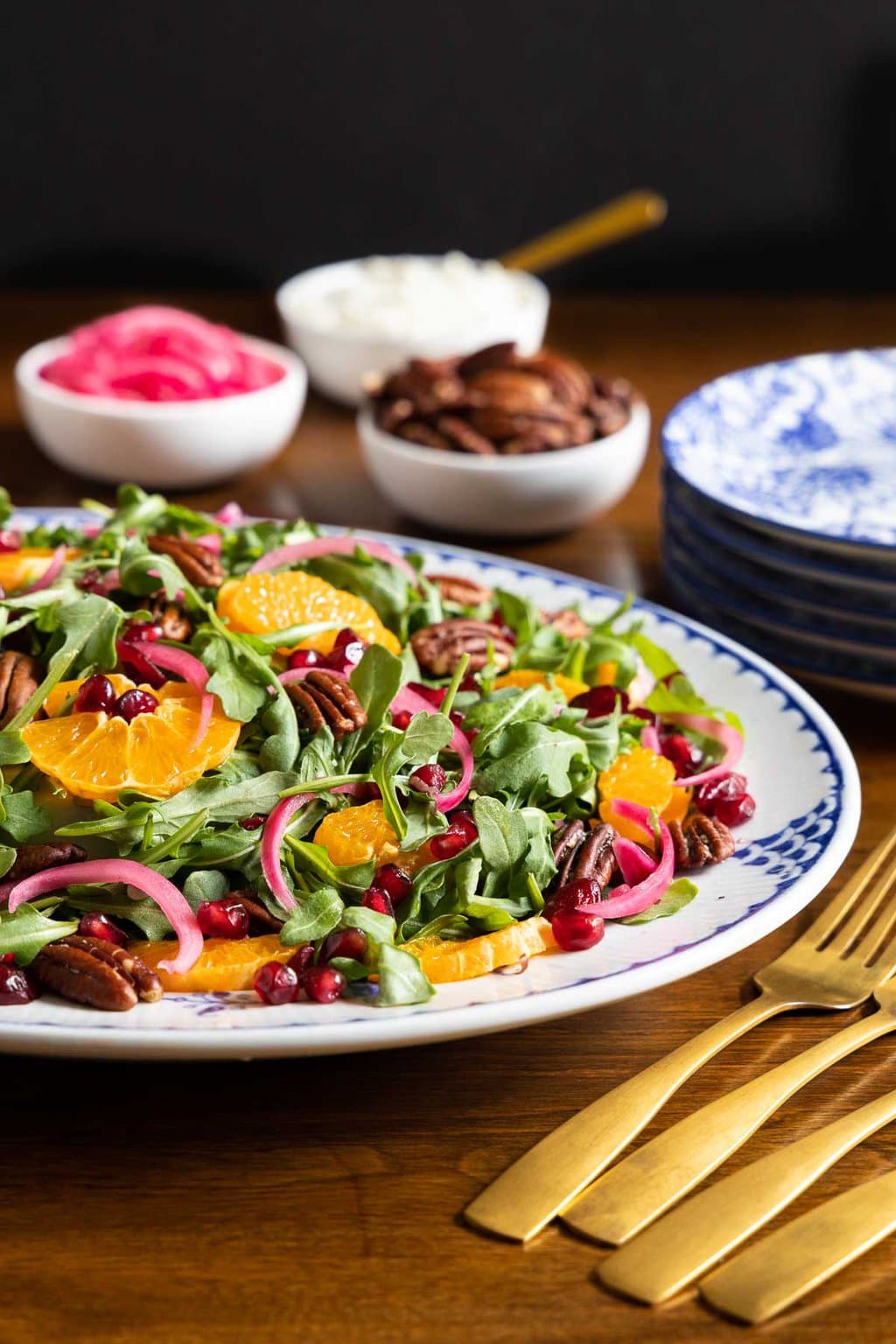 Horizontal photo of a Clementine Arugula Cranberry Salad on a blue and white patterned platter surrounded by bowls of garnishes for the salad.