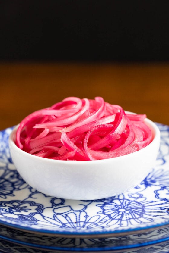 Vertical closeup photo of a bowl of Clementine-Pickled Red Onions on a stack of blue and white patterned serving plates.