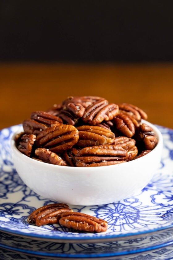 Vertical closeup photo of a bowl of Butter-Roasted Pecans on a stack of blue and white patterned serving plates.