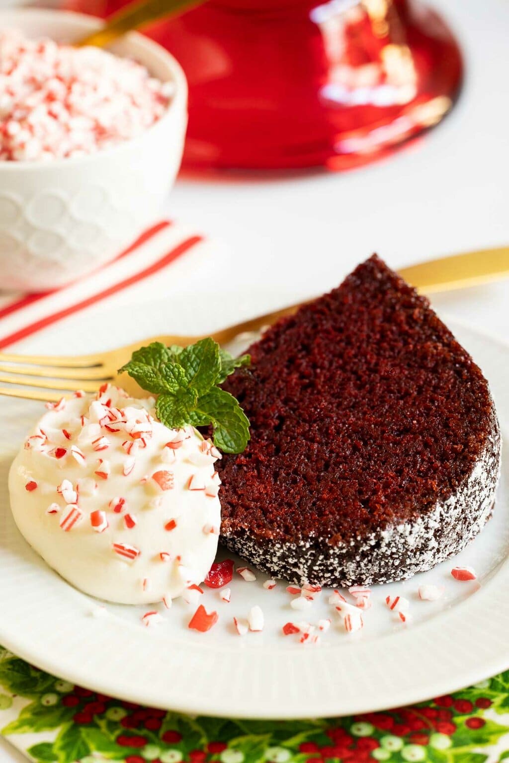 Vertical photo of a slice of Ridiculously Easy Peppermint-Glazed Red Velvet Bundt Cake with a dollop of whipped cream, a sprig of fresh mint and crushed peppermint candy cane pieces.