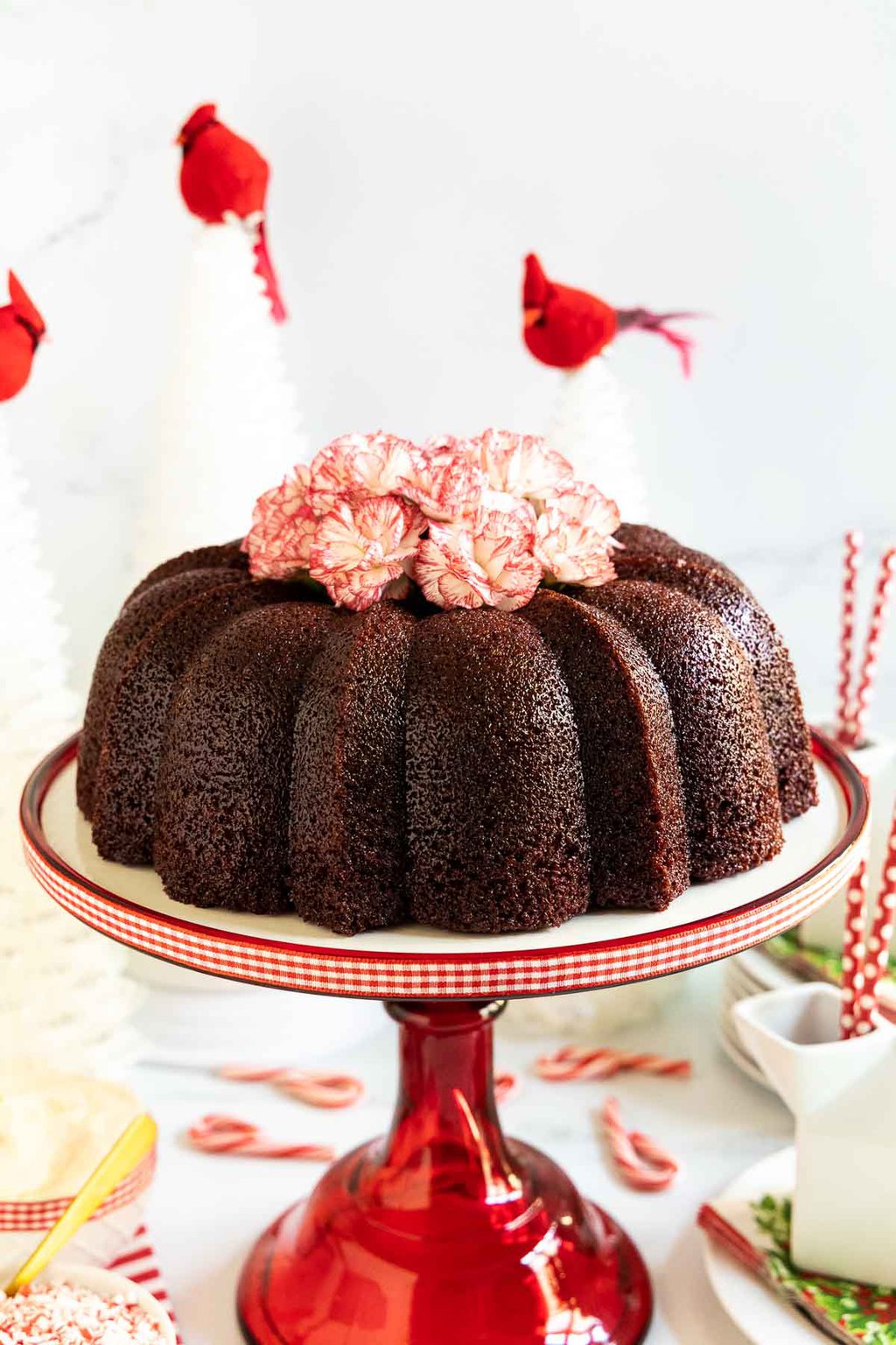 Vertical photo of a Ridiculously Easy Peppermint-Glazed Red Velvet Bundt Cake surrounded by cartons of milk and candy canes and decorated with red and white carnations.