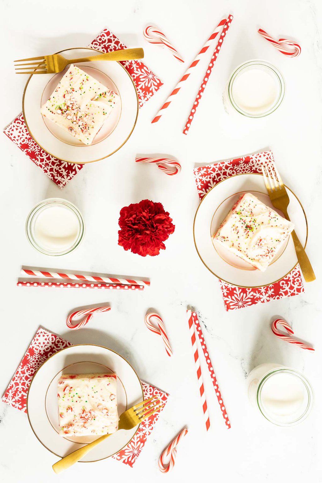 Vertical overhead photo of three slices of Ridiculously Easy Candy Cane Sheet Cake on individual white and gold serving plates surrounded by candy canes and Christmas straws.