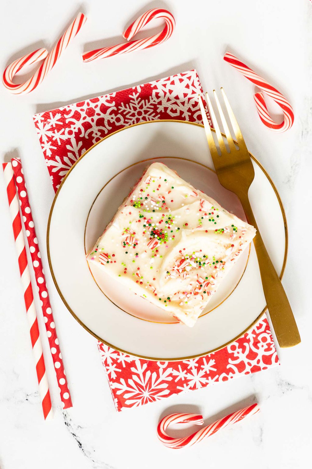 Overhead vertical photo of a slice of Ridiculously Easy Candy Cane Sheet Cake on a white and gold serving plate with colorful Christmas candy canes and straws.