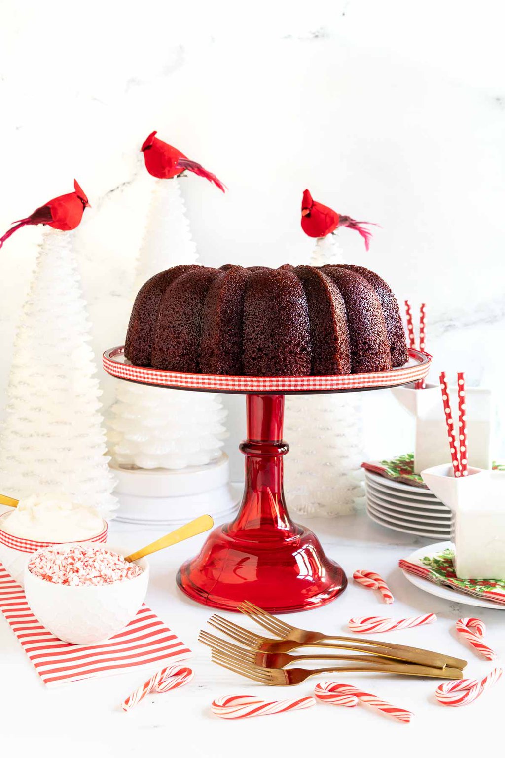 Vertical photo of a Ridiculously Easy Peppermint-Glazed Red Velvet Bundt Cake on a red glass pedestal cake stand surrounded by cardinals and candy canes.