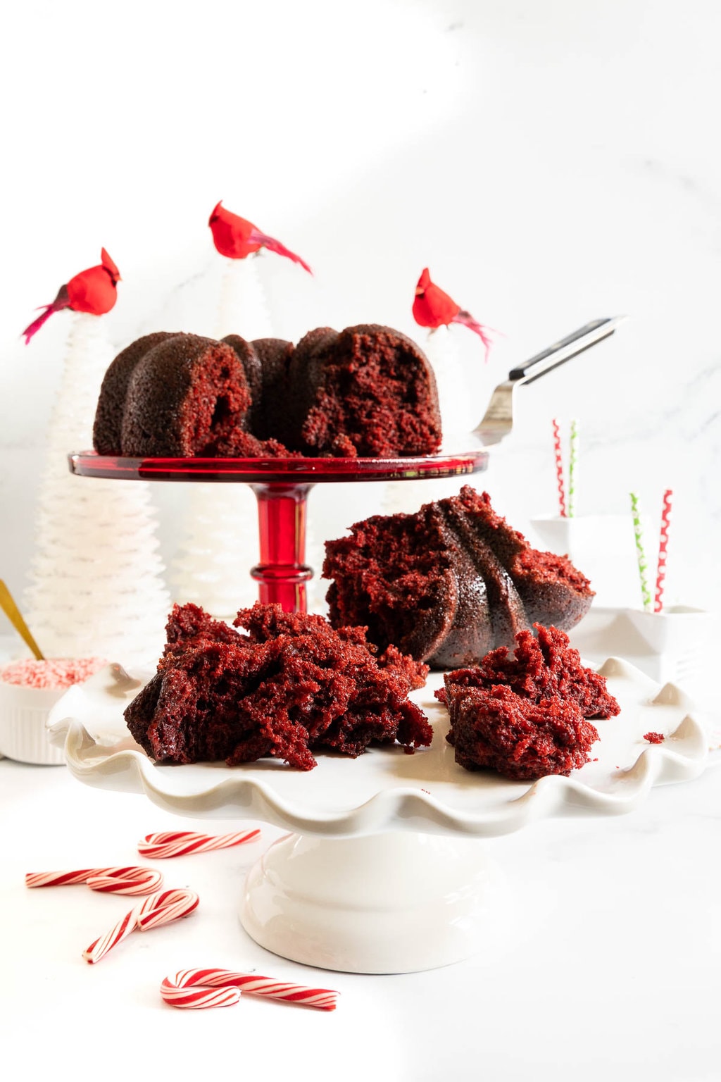 Vertical photo of broken pieces of a Ridiculously Easy Peppermint-Glazed Red Velvet Bundt Cake on two pedestal cake stands.