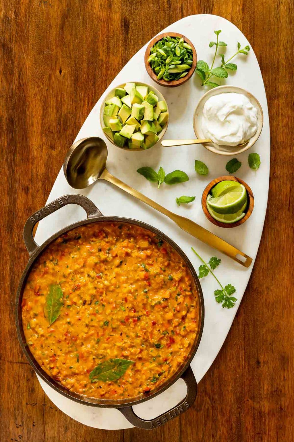 Vertical overhead photo of a cast iron pot of Curried Red Lentil Coconut Soup with small bowls of diced avocados, lime wedges, sliced green onions and fresh basil and cilantro leaves.