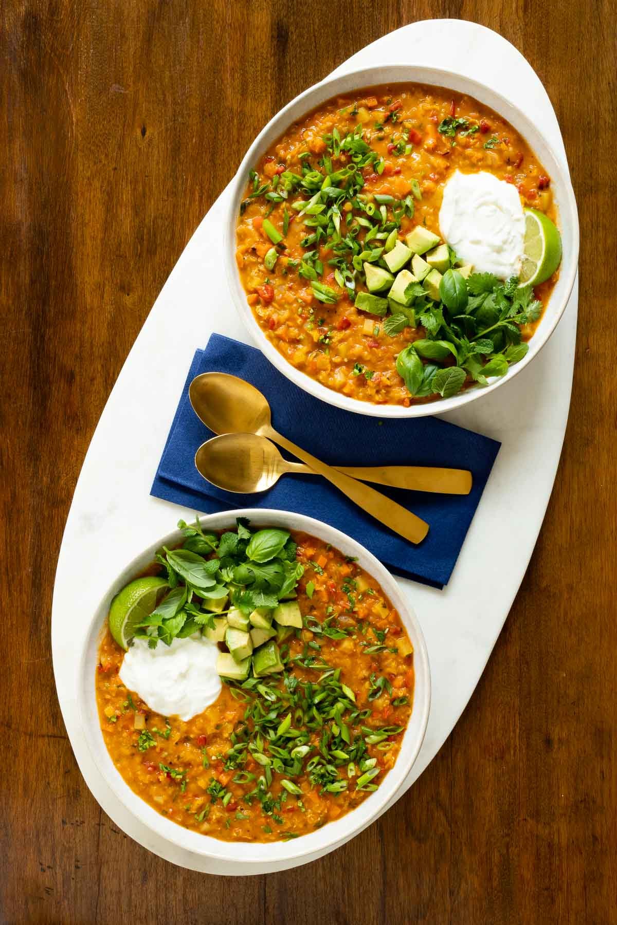 Vertical overhead photo of two white serving bowls of Curried Red Lentil Coconut Soup garnished with avocados, sliced green onions, lime wedges, and fresh cilantro and basil leaves.