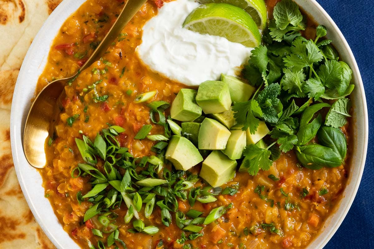 Horizontal closeup photo of a serving bowl of Curried Red Lentil Coconut Soup garnished with lime wedges, cilantro, basil avocados and sliced green onions.
